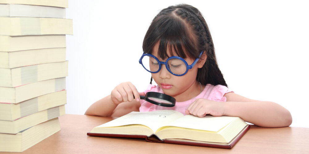 A young girl of Asian descent studies a book with a magnifying glass. She's wearing comically large glasses.