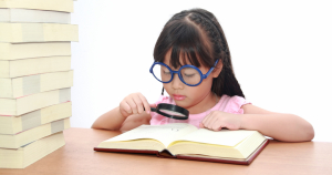 A young girl of Asian descent studies a book with a magnifying glass. She's wearing comically large glasses.